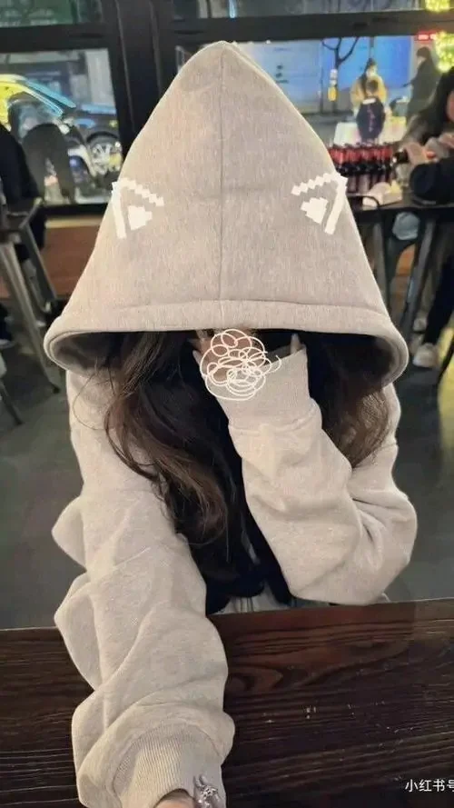 Mũ trùm tai mèo che gần hết gương mặt, cô gái ngồi trong quán, áo kem rộng và tay chống cằm tạo nét đáng yêu