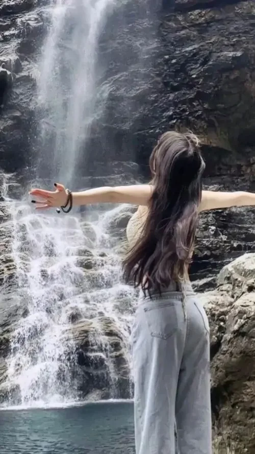 Dòng thác trắng phía sau làm mái tóc dài mềm hơn, cô gái quay lưng vươn tay nhẹ, váy xám tạo dáng thướt tha giữa thiên nhiên