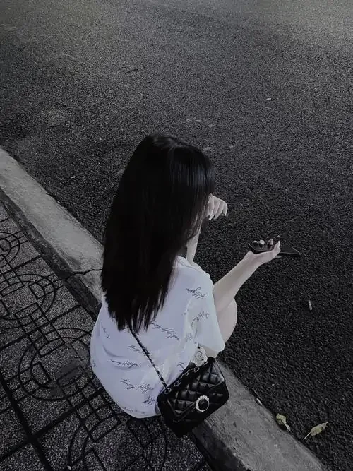 Góc nhìn từ trên cao bắt dáng ngồi bên vỉa hè, tóc đen xõa ngang vai che nửa mặt, áo trắng và túi nhỏ rất đời thường
