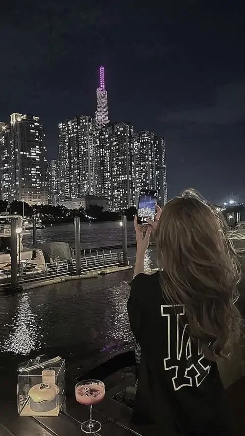 Ánh đêm thành phố phía xa làm bóng lưng cô gái thêm lặng, áo đen in chữ và tóc nâu dài che gần hết khuôn mặt