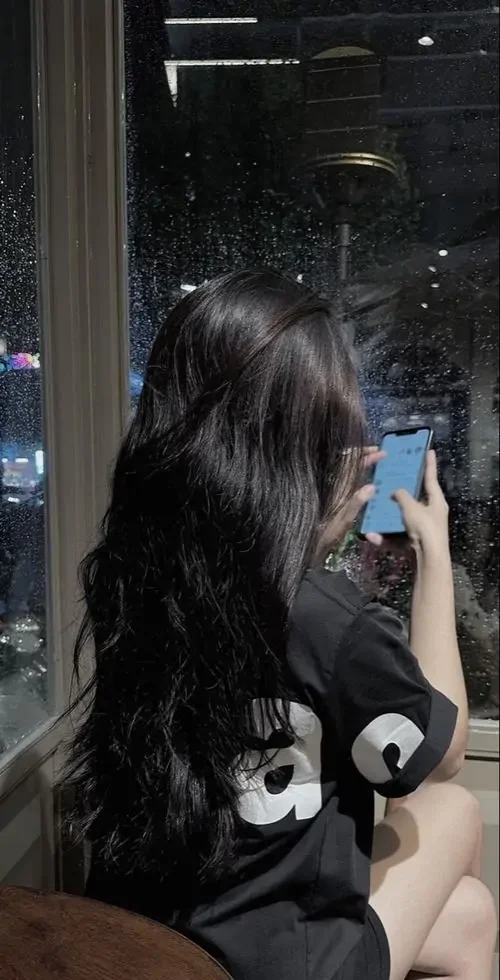 Trước ô kính mờ phản chiếu ánh đêm, cô gái tóc dài cầm điện thoại che mặt, áo đen tạo nét riêng tư và hơi lạnh